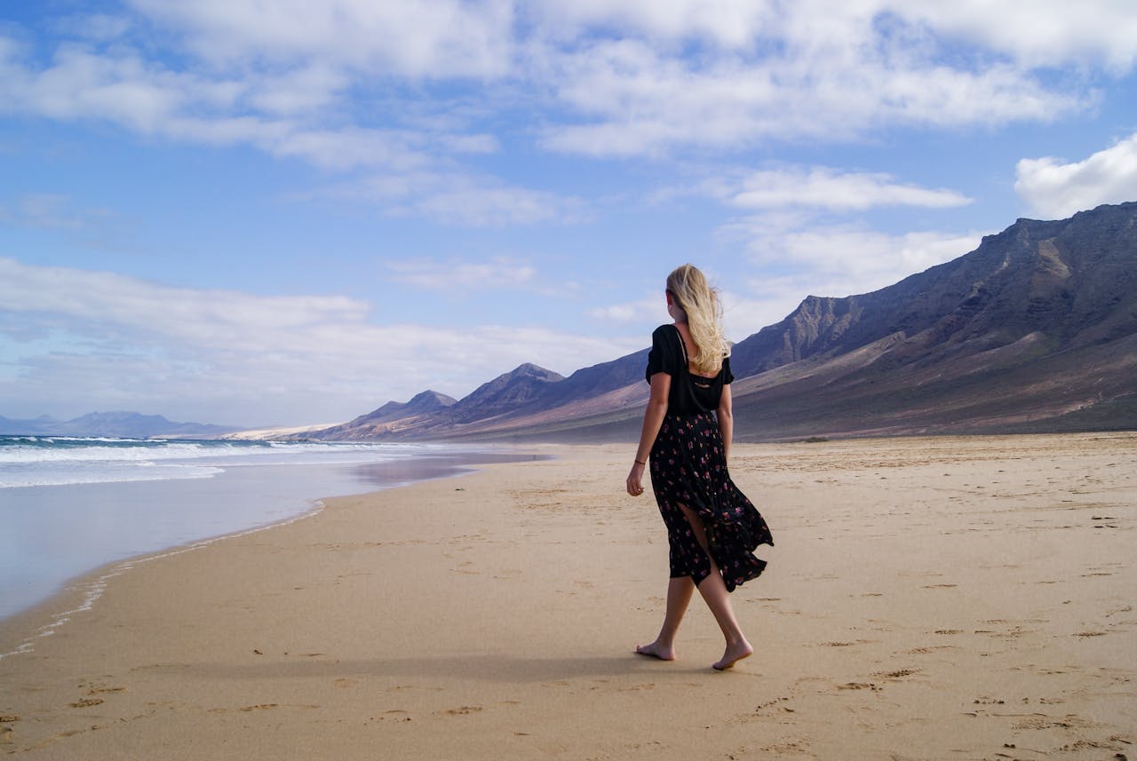 Woman with long blonde hair strolls on Cofete Beach, Spain, showcasing fashion in a serene beach setting.